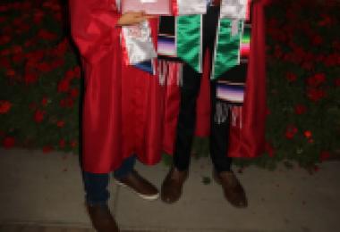 Two male graduates in red caps and gowns pose with diplomas at night, surrounded by flowers.