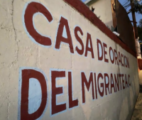 Painted wall sign reading 'Casa de Oracion del Migrante' with red trim, sunlit