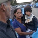 Woman holding young child amid blue tent encampment, anxious expression; blurred man nearby.